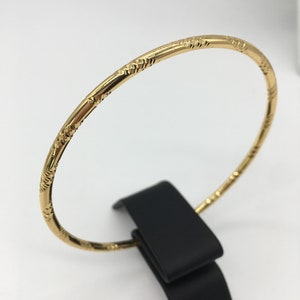 May include: A gold bangle bracelet with a textured design. The bracelet is on a black display stand.