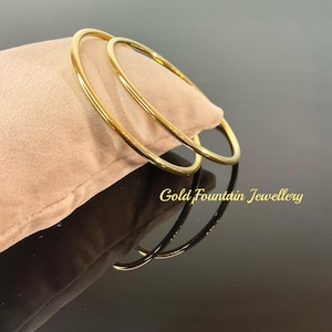 May include: Two gold bangle bracelets on a tan velvet cushion. The bracelets are simple and smooth, with a polished finish. The text "Gold Fountain Jewellery" is visible below the bracelets.