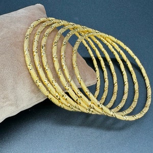 May include: A set of seven gold-coloured bangles with a textured design. The bangles are stacked on top of each other.