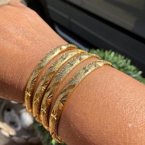 May include: Six gold bangles, each with a textured surface and a repeating starburst pattern. The bangles are stacked on a wrist, highlighting their design. The jewelry is displayed in natural light, showcasing the gold's shine.