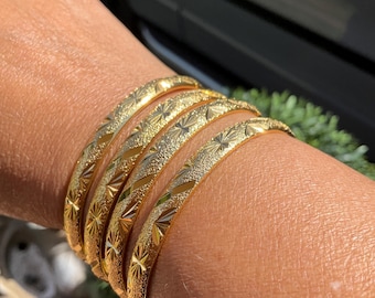 Peut inclure: Ensemble de six bracelets dorés. Chaque bracelet présente une surface texturée avec un motif d'éclat d'étoile répété. Les bracelets sont empilés sur un poignet, mettant en valeur leur design et leur éclat. Bijoux en lumière naturelle.