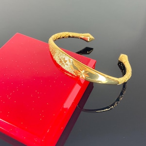May include: A gold-coloured open cuff bracelet featuring a polished centre section with two engraved star-like floral motifs, flanked by textured, matte-finished bands that taper towards pointed, smooth, and solid gold-coloured terminal ends.