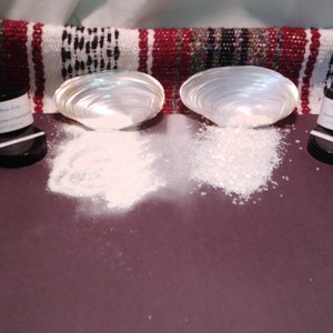 May include: Two iridescent seashell halves flank piles of white powder and crystals. Small black jars with white labels are on either side. The background features a red, white, and black woven textile.