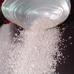 May include: A white seashell filled with iridescent white crushed seashells, spilling out onto a dark surface.