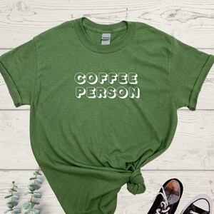 May include: A green t-shirt with the text "COFFEE PERSON" in white, 3D-style lettering. The shirt is knotted and laid on a wooden surface with a pair of black Converse sneakers.