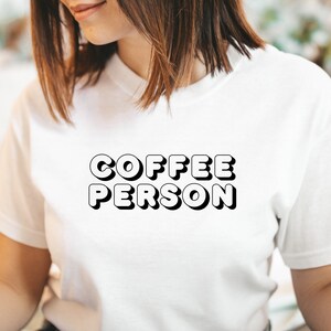 May include: White t-shirt with the text "COFFEE PERSON" in black, 3D-style lettering.