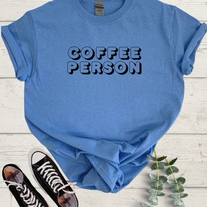 May include: A light blue t-shirt with the text "COFFEE PERSON" in black, 3D-style lettering. The shirt is folded and displayed on a white wooden surface with a pair of black sneakers and a sprig of green foliage.