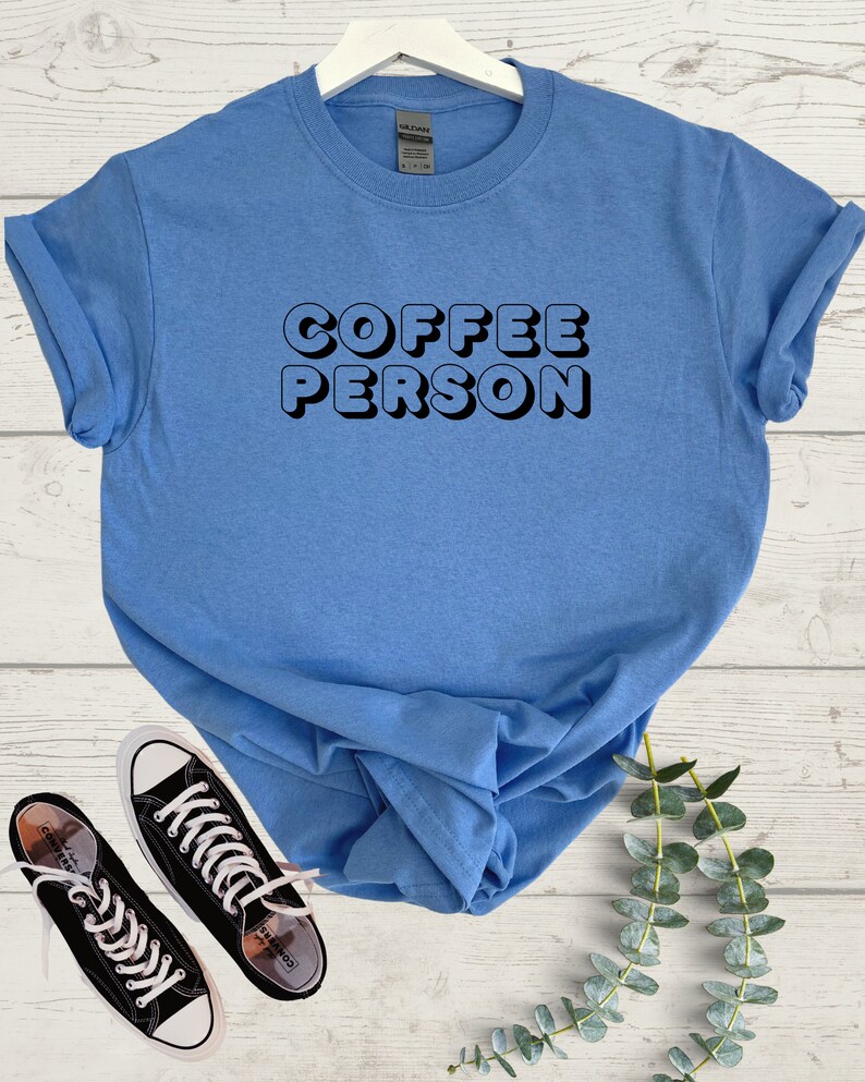 May include: A light blue t-shirt with the text "COFFEE PERSON" in black, 3D-style lettering. The shirt is folded and displayed on a white wooden surface with a pair of black sneakers and a sprig of green foliage.
