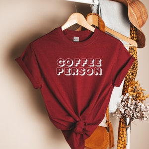May include: A maroon t-shirt with the text "COFFEE PERSON" in white outlined letters. The shirt is knotted at the bottom and hanging on a wooden hanger.