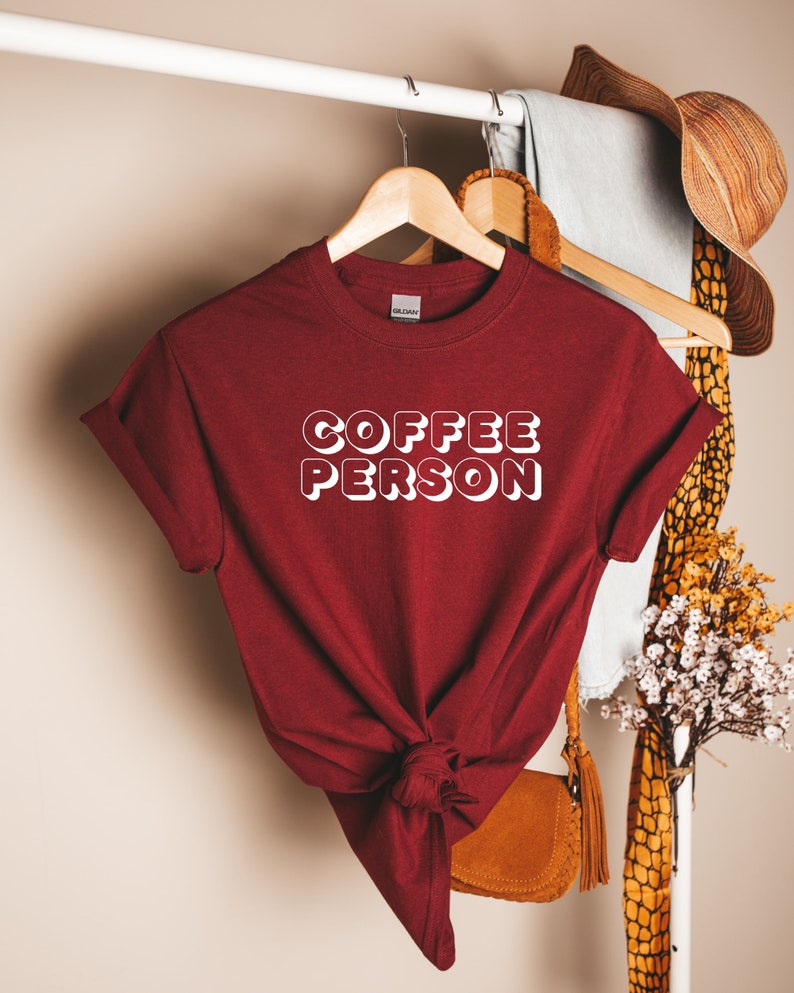 May include: A maroon t-shirt with the text "COFFEE PERSON" in white outlined letters. The shirt is knotted at the bottom and hanging on a wooden hanger.
