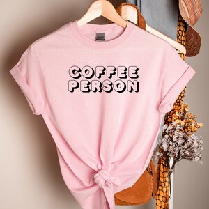 May include: Pink t-shirt with black text that reads "COFFEE PERSON" in a retro style font.
