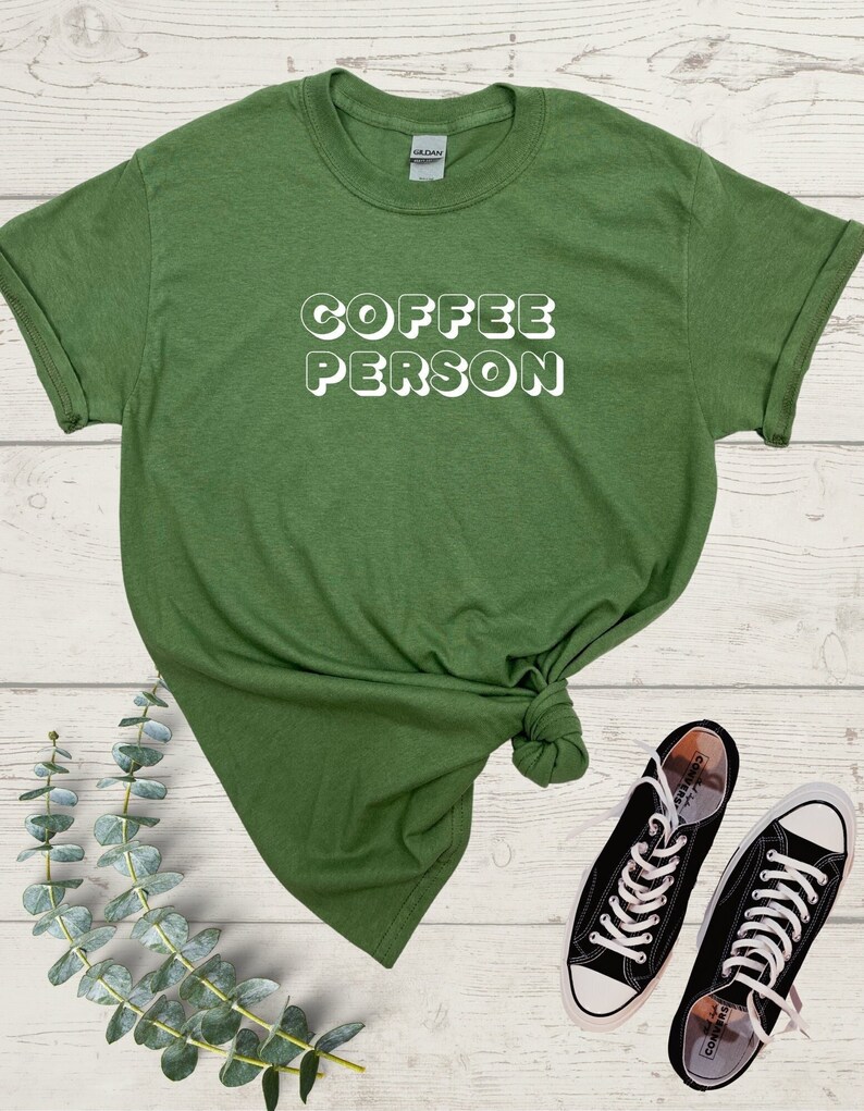 May include: A green t-shirt with the text "COFFEE PERSON" in white, 3D-style lettering. The shirt is knotted and laid on a wooden surface with a pair of black Converse sneakers.