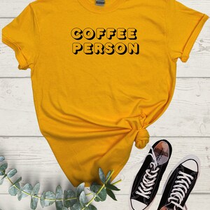 May include: A yellow t-shirt with the text "COFFEE PERSON" in black outlined letters. The shirt is on a white wooden surface with a sprig of eucalyptus leaves and two black Converse sneakers.