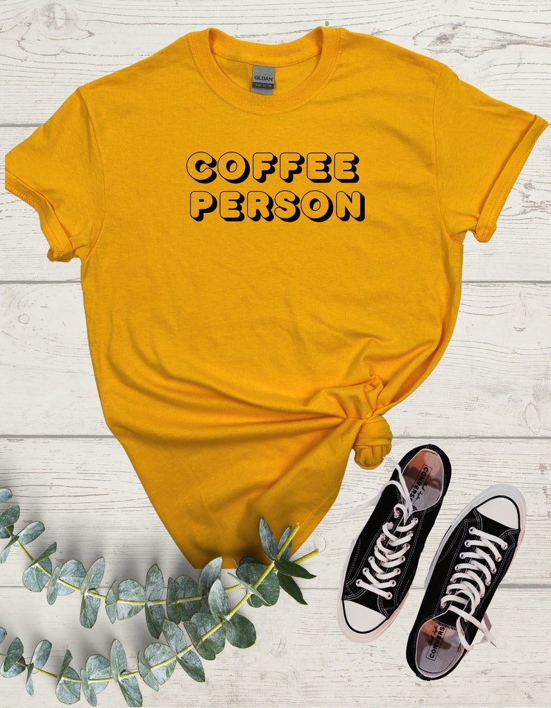 May include: A yellow t-shirt with the text "COFFEE PERSON" in black outlined letters. The shirt is on a white wooden surface with a sprig of eucalyptus leaves and two black Converse sneakers.