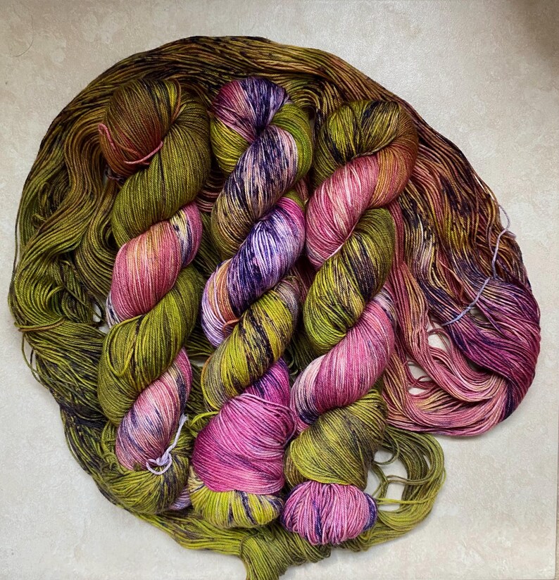 Op de afbeelding: Vier bollen handgeverfde garen in tinten van groen, roze en paars. Het garen is in een cirkel gerangschikt, met de uiteinden van de bollen die elkaar overlappen.