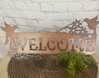 May include: A metal welcome sign with a rustic brown finish. The sign features the word "WELCOME" in a decorative font and is adorned with two hummingbirds and floral accents.