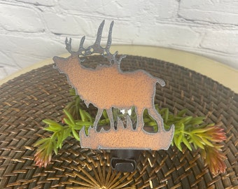 May include: A metal silhouette of an elk with a light bulb in the center. The elk is a rusty brown color and is standing on a green base. The silhouette is attached to a black plug.