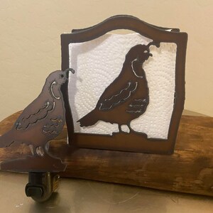 May include: A brown metal napkin holder with a quail silhouette design. The holder is sitting on a wooden surface. There is a smaller quail silhouette on the left side of the holder.