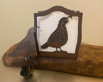 May include: A brown metal napkin holder with a quail silhouette design. The holder is sitting on a wooden surface. There is a smaller quail silhouette on the left side of the holder.