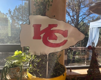 May include: A white arrowhead-shaped sign with the letters "KC" in red, with a white background and red outline. The sign is on a white stick and is in front of a window.
