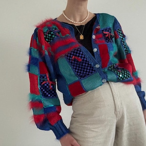 Könnte beinhalten: Ein farbenfroher Patchwork-Cardigan mit V-Ausschnitt, der rote, blaue und türkisfarbene Quadrate kombiniert. Der Pullover hat einen flauschigen roten Besatz und Pailletten-Akzente. Die Ärmel sind lang und der Cardigan ist geknöpft.