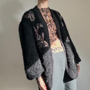 Könnte beinhalten: Ein schwarzer Cardigan mit offenem Schnitt und strukturiertem, flauschigem Aussehen. Der Cardigan hat gestickte Blumen- und Kaninchenmuster in Lila-, Grün- und Grautönen. Die Kanten sind mit dunklerem Strickmaterial verziert. Der Cardigan wird über einem gemusterten Oberteil und einer gestreiften Hose getragen.