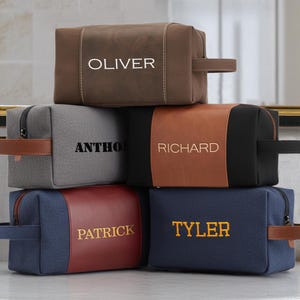 May include: A stack of personalized toiletry bags in various colors, including brown, gray, navy, and black. Each bag features a different name embroidered in a contrasting color. The bags have handles and zippers.