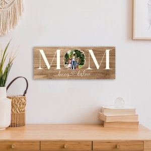 Puede incluir: Un letrero de madera con la palabra "MOM" en letras blancas con una corona de flores alrededor de la "O". Una foto de una mujer y dos ni&ntilde;os est&aacute; dentro de la corona. El texto "denny & darleen" est&aacute; debajo de la palabra "MOM".