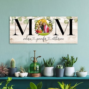 Puede incluir: Un letrero de madera con la palabra "MOM" en letras negras grandes. Las letras est&aacute;n rodeadas de hojas verdes y un c&iacute;rculo con una foto de tres personas dentro. El texto "valerie jennifer catherine" est&aacute; escrito debajo de la palabra "MOM".