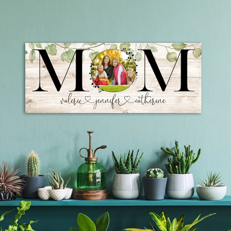 Puede incluir: Un letrero de madera con la palabra "MOM" en letras negras grandes. Las letras est&aacute;n rodeadas de hojas verdes y un c&iacute;rculo con una foto de tres personas dentro. El texto "valerie jennifer catherine" est&aacute; escrito debajo de la palabra "MOM".