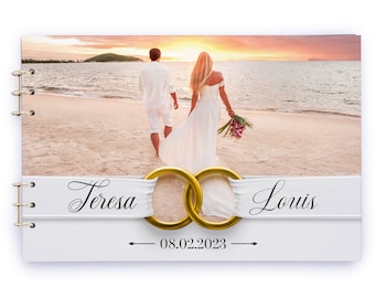 May include: A wedding guest book with a photo of a couple on a beach at sunset. The book has a white ribbon with the names "Teresa" and "Louis" and the date "08.02.2023" printed on it.