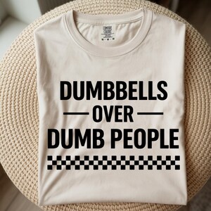 May include: A beige t-shirt with the text "DUMBBELLS OVER DUMB PEOPLE" in bold black letters. A black and white checkered pattern is at the bottom of the text. The shirt is folded neatly.