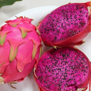 Puede incluir: Tres frutas del dragón, una entera y dos cortadas por la mitad, en un plato blanco. La fruta es rosa con escamas verdes y tiene una pulpa morada con semillas negras.