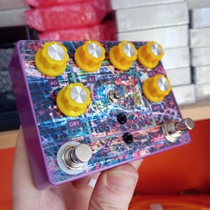 Può includere: Un pedale per effetti viola con un design astratto colorato. Presenta sei manopole gialle con coperchi argentati, due interruttori a pedale e un interruttore a levetta. Le etichette "A0", "A1", "A2", "DRY" e "WET" sono presenti.