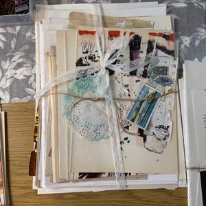 Könnte beinhalten: Ein Stapel Vintage-Papier-Ephemera, gebunden mit Spitzenband und Bindfaden. Das Bündel enthält verschiedene Papiersorten, eine Deckchen und kleine Kunstwerke. Die Papiere sind cremefarben, einige mit farbigen Akzenten. Eine kleine blau-weiße Karte ist sichtbar.