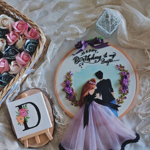 Peut inclure: Art de broderie en forme de cercle représentant un couple dans une pose romantique. La femme porte une robe violette avec un ruban noir et l'homme porte un costume noir. L'arrière-plan est un ciel bleu clair avec des fleurs violettes. Le texte "Happy Birthday to my Buji..." est écrit en cursive noire.