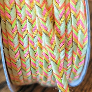 Puede incluir: Primer plano de un carrete de ribete trenzado verde neón, rosa y amarillo con hilo plateado brillante tejido en todo.