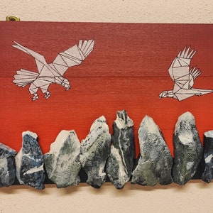Può includere: Un'opera d'arte murale in legno con uno sfondo sfumato rosso e arancione. Due aquile geometriche bianche volano in direzioni opposte. La parte inferiore del pezzo presenta una fila di rocce grigie.