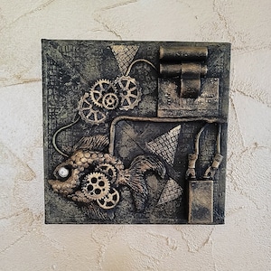 Può includere: Un'opera d'arte murale quadrata, di colore verde scuro e dorato, che presenta un pesce stilizzato con ingranaggi e altri accenti metallici. Il pesce è circondato da forme e motivi geometrici.