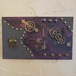 Può includere: Un portachiavi in legno viola con ingranaggi metallici, chiavi e gufi. Il portachiavi ha quattro ganci per appendere le chiavi.