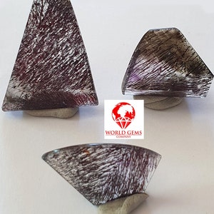 May include: Three clear quartz crystals with black inclusions, also known as rutilated quartz, in various sizes and shapes. The crystals are displayed on a white surface.