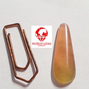 May include: A teardrop-shaped, translucent yellow gemstone with a slight pink hue. The gemstone has a smooth, polished surface and is set against a white background.