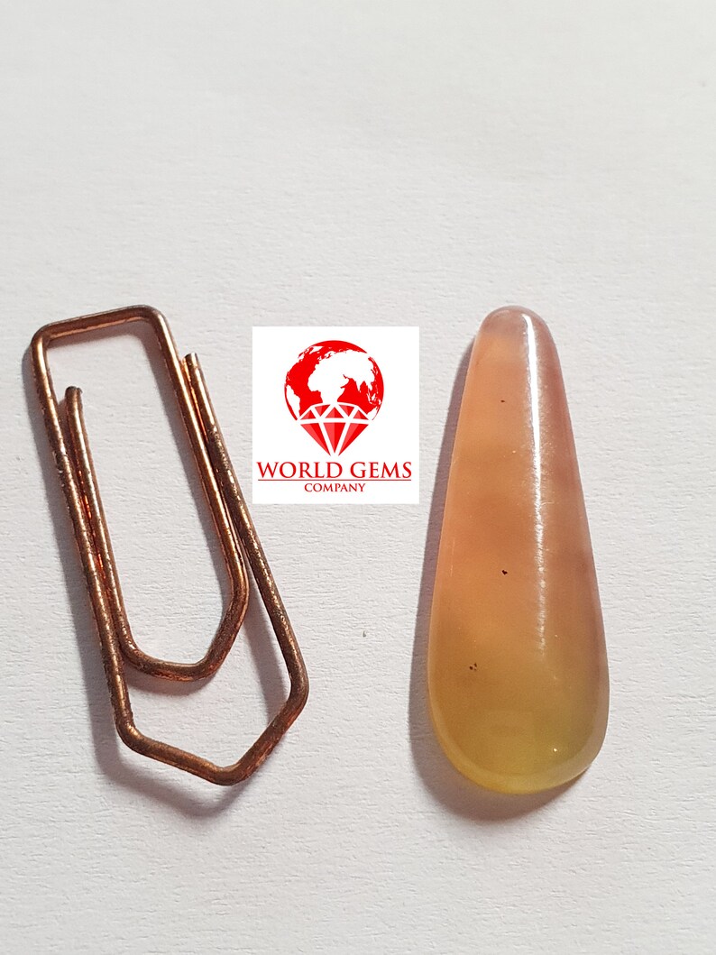 May include: A teardrop-shaped, translucent yellow gemstone with a slight pink hue. The gemstone has a smooth, polished surface and is set against a white background.