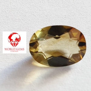 Puede incluir: Una piedra preciosa ovalada de color marrón dorado con un patrón en forma de estrella en su interior. La piedra preciosa está sobre una superficie blanca.