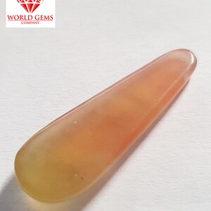 May include: A long, thin, teardrop-shaped gemstone with a pale orange and yellow colour. The gemstone is translucent and has a smooth, polished surface.