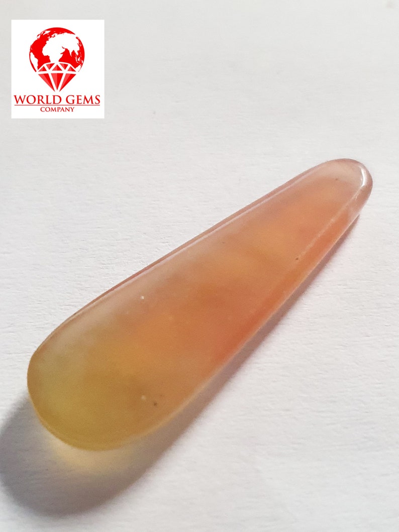 May include: A long, thin, teardrop-shaped gemstone with a pale orange and yellow colour. The gemstone is translucent and has a smooth, polished surface.