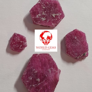 Op de afbeelding: Vier ruwe, rode robijn edelstenen in verschillende maten en vormen.  WORLD GEMS COMPANY is gedrukt in witte tekst op een rode achtergrond.