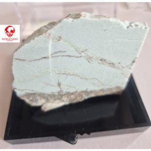 May include: A light blue gemstone with brown veining, displayed on a black stand. The stone has a rough, natural texture and is irregularly shaped. The World Gems Company logo is visible in the upper left corner.