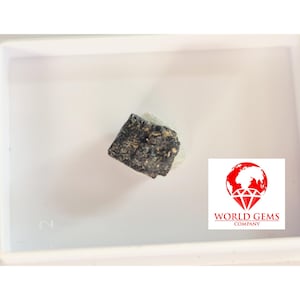 May include: A dark, rough-textured gemstone, possibly a mineral specimen, set against a white background. The image includes a logo with a globe and the text "WORLD GEMS COMPANY".