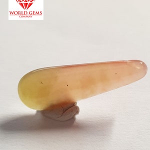 May include: A long, smooth, light pink and yellow gemstone with a slightly pointed end. The gemstone is sitting on a small, grey, textured base.